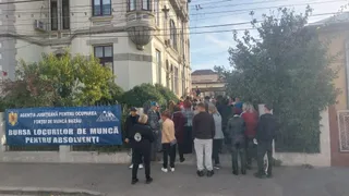 Un grup de persoane stă în picioare pe un drum, lângă un banner albastru și în fața unei clădiri mari cu ferestre și vegetație.