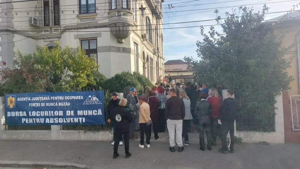 Un grup de persoane stă în picioare pe un drum, lângă un banner albastru și în fața unei clădiri mari cu ferestre și vegetație.