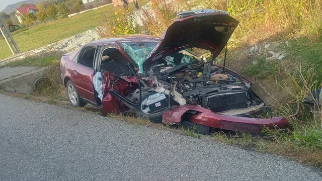 Un autoturism roșu cu capota și partea frontală deformată stă pe marginea unui drum asfaltat lângă un taluz cu iarbă.