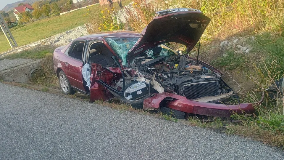Un autoturism roșu cu capota și partea frontală deformată stă pe marginea unui drum asfaltat lângă un taluz cu iarbă.