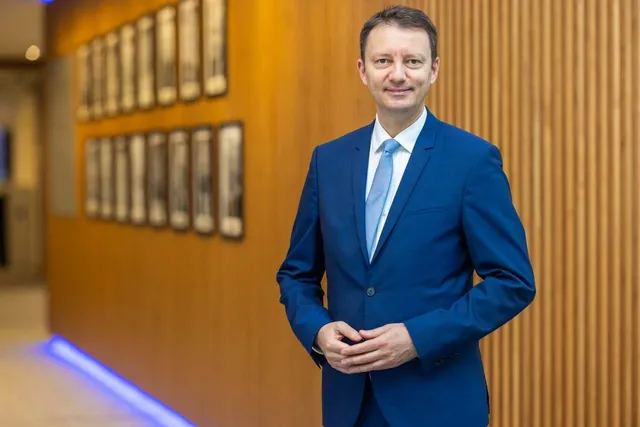 Un bărbat cu păr scurt, purtând un costum albastru și o cămașă albă cu cravată albastră, stă în picioare în fața unui perete din lemn.