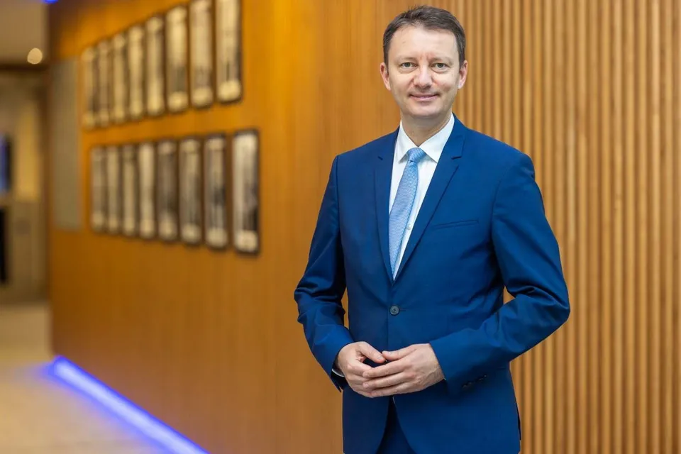 Un bărbat cu păr scurt, purtând un costum albastru și o cămașă albă cu cravată albastră, stă în picioare în fața unui perete din lemn.