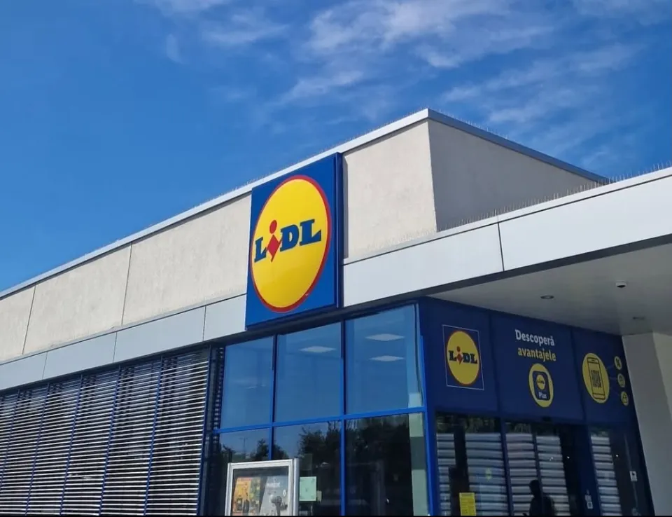 Fațada unei clădiri comerciale cu panouri de sticlă albastră și un panou publicitar galben cu un logo pe fundal albastru.