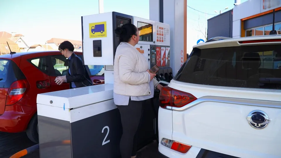 O femeie cu o geantă mică stă lângă o pompă de benzină lângă un autoturism alb, în timp ce un bărbat stă lângă o mașină roșie.