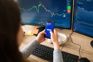 O persoană ține un smartphone cu un ecran albastru în fața a două monitoare care afișează grafice financiare pe un birou cu tastatură și mouse.