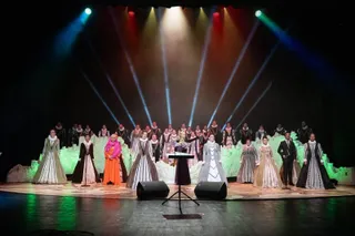 Un grup de persoane în costume lungi și colorate stă pe o scenă iluminată de proiectoare colorate sub un fundal întunecat.