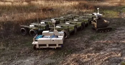 Mai multe vehicule robotizate cu roți și șenile sunt parcate pe un teren cu iarbă uscată și pământ.