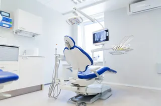 Un scaun de stomatologie albastru și alb este poziționat într-o cameră medicală cu un monitor și un aparat de iluminat suspendate.