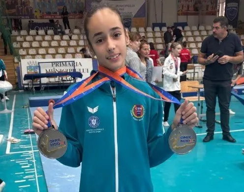 O tânără poartă un tricou albastru și prezintă două medalii argintii cu pangalii portocalii și albastre într-o sală de sport cu oameni în fundal.