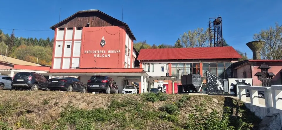 O clădire cu fațadă roșie și albă, pe care scrie text, este situată lângă un ansamblu de construcții cu acoperișuri roșii și mașini parcate în față.
