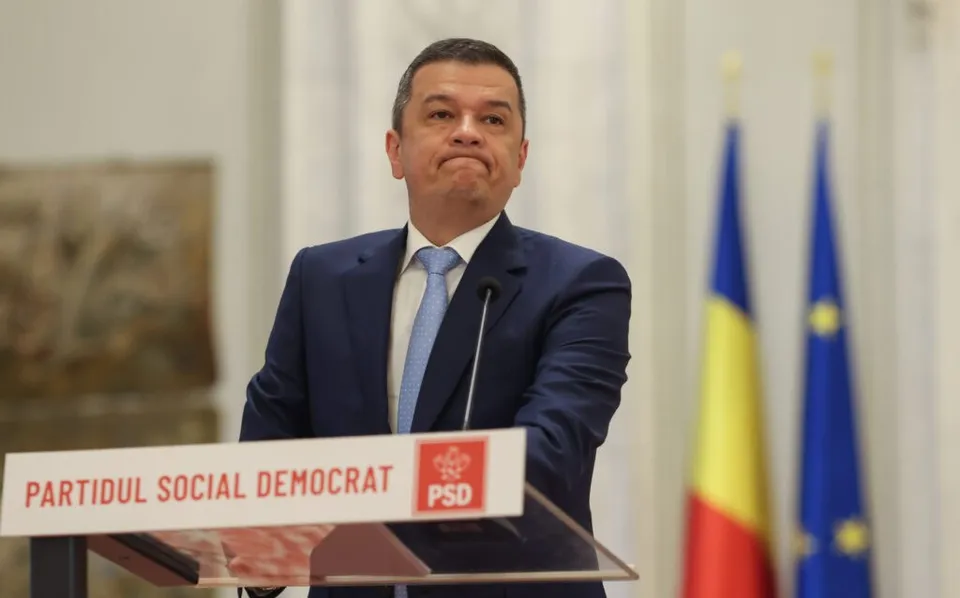Un bărbat într-un costum albastru și cravată deschisă stă în spatele unui podium alb cu inscripția PARTIDUL SOCIAL DEMOCRAT.