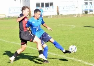 Un jucător în echipament albastru lovește un balon pe un teren de fotbal în prezența unui adversar îmbrăcat în negru și roșu.