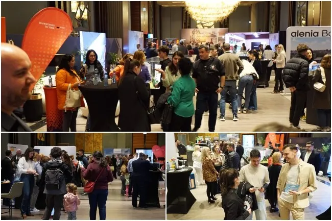 Oamenii stau în grupuri într-o sală de evenimente cu mese negre, standuri de expoziție și un candelabru suspendat în fundal.