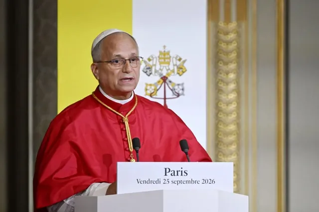 Un preot în costum roșu cu guler alb și ochelari vorbește la un podium alb pe care scrie Paris, cu un fundal galben și papal.