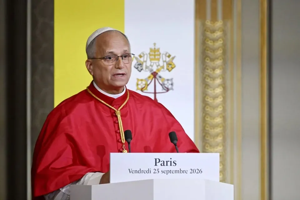 Un preot în costum roșu cu guler alb și ochelari vorbește la un podium alb pe care scrie Paris, cu un fundal galben și papal.