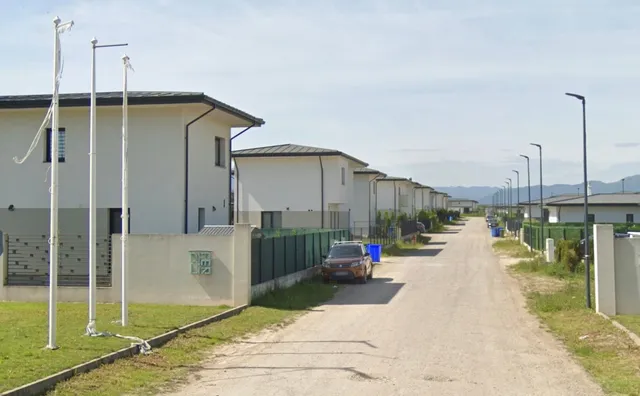 O serie de case albe cu acoperișuri înclinat sunt situate de-a lungul unui drum de țărână sub un cer înnorat.