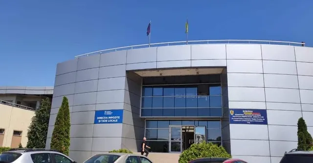 O clădire modernă cu fațadă gri metalică, ferestre mari de sticlă și două steaguri pe acoperiș, aflată sub un cer senin.