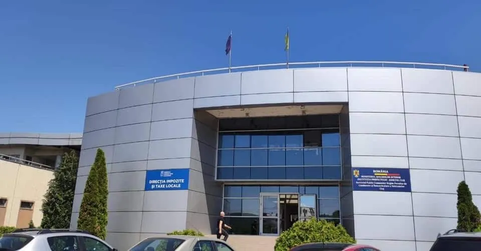 O clădire modernă cu fațadă gri metalică, ferestre mari de sticlă și două steaguri pe acoperiș, aflată sub un cer senin.
