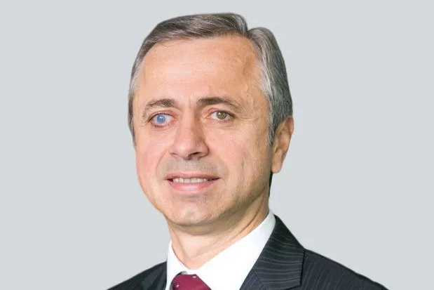 Un bărbat cu părul cărunt și ochi de culori diferite poartă o cămașă albă, o cravată roșie și un sacou în dungi fine.