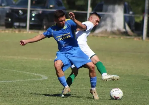 Un jucător în echipament albastru și unul în echipament alb se confruntă pe un teren de fotbal lângă un balon.
