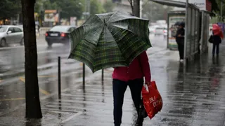 O persoană cu o umbrelă în model tartan merge pe un trotuar umed lângă un autobuz, purtând o sacoșă roșie.