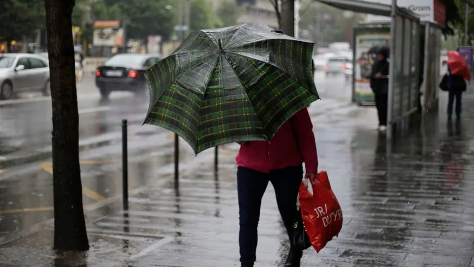 O persoană cu o umbrelă în model tartan merge pe un trotuar umed lângă un autobuz, purtând o sacoșă roșie.