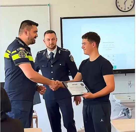 Un bărbat în uniformă de polițist strânge mâna unui tânăr care ține o diplomă într-o încăpere cu un ecran interactiv în fundal.