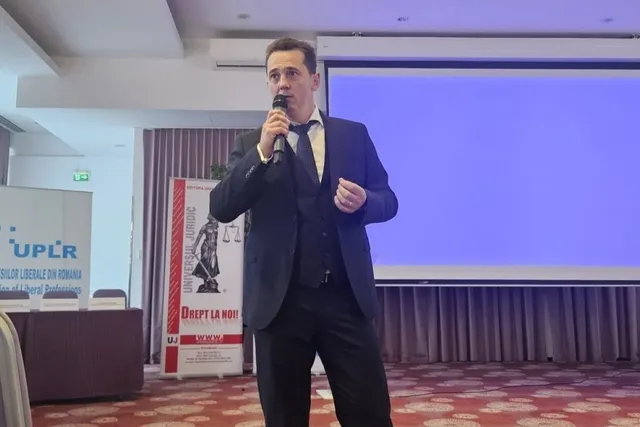 Un bărbat într-un costum închis la culoare ține un microfon și stă în fața unui ecran de proiecție albastru într-o încăpere.