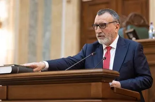 Un bărbat cu ochelari și barbă scurtă, purtând costum închis și cravată roșie, stă în spatele unui podium de lemn întinsând mâna pe o carte.