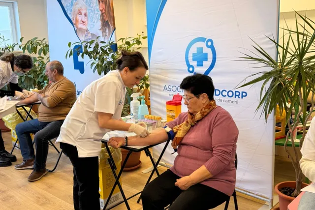 O femeie în halat alb consultă brațul unei femei așezate pe un scaun într-un spațiu cu plante și un panou informativ.