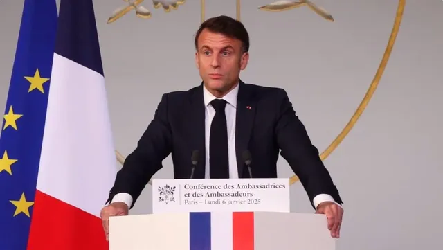 Un bărbat într-un costum închis și cravată neagră stă în spatele unui podium alb lângă un steag european și un steag francez.