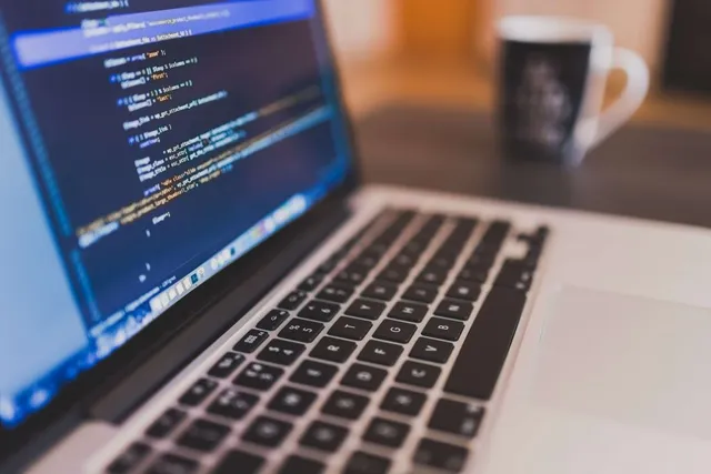 Un laptop deschis afișează linii de cod pe un ecran albastru, fiind plasat pe o masă lângă o cană de cafea neagră.