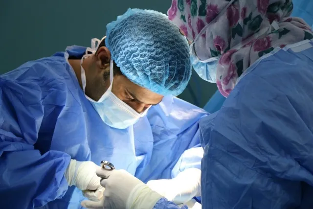 Două persoane purtând halate și căciuli de protecție albastre efectuează o procedură medicală sub o lumină intensă.