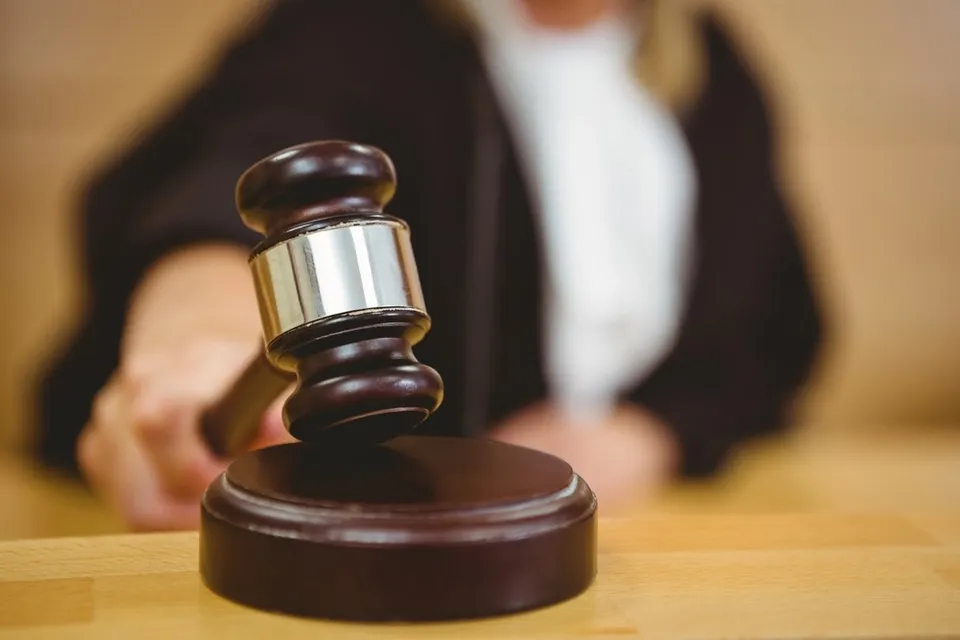 Un ciocan de judecător din lemn cu detalii metalice este așezat pe o masă de lemn în fața unei persoane purtând o robă neagră.