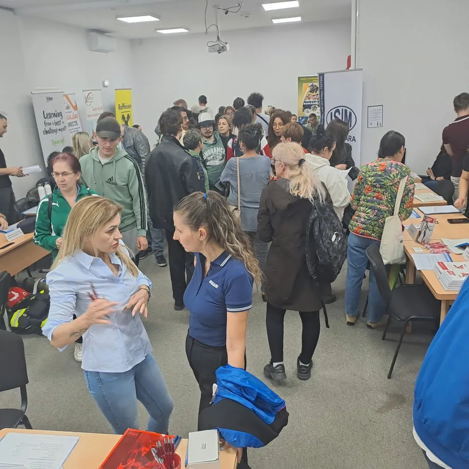 O grupă de oameni stă în picioare într-o încăpere cu mese, având diverse materiale de lucru și panouri informative în fundal.