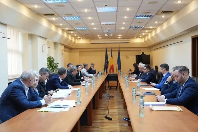 O grupă de persoane stă la mese lungi de conferință într-o încăpere cu tavanul iluminat de panouri rectangulare, având steaguri în fundal.