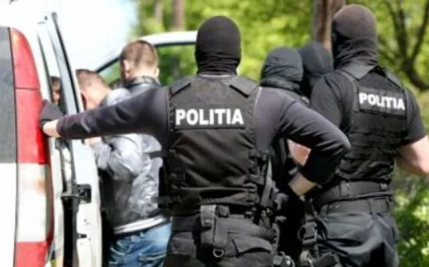 Patru polițiști în uniformă neagră cu inscripția "POLITIA" și măști faciale întunecate stau lângă o mașină albă lângă un grup de persoane.