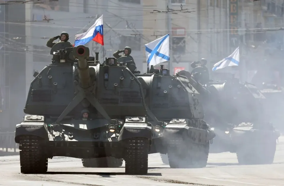 Trei vehicule militare blindate cu tunuri mari circulă pe un drum, în timp ce soldați stau în partea superioară și flutură steaguri.