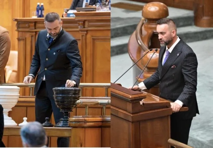 Un bărbat cu barbă poartă costum și stă în spatele unui pupitru de lemn, lângă un ciocan de judecată de dimensiuni mari.