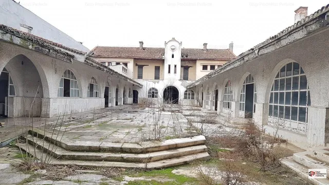 O curte pavată cu pietre și trepte de beton este înconjurată de clădiri albe cu ferestre și arcade mari sub un cer deschis.