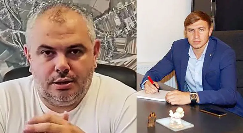 Un bărbat cu barbă poartă un tricou alb, iar lângă el, un alt bărbat cu sacou albastru stă la o masă cu un caiet și un pix.