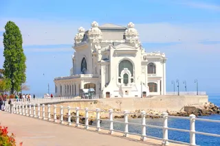 O clădire albă de arhitectură clasică se află pe malul mării, lângă un drum pietonal cu balustradă albă sub un cer senin.