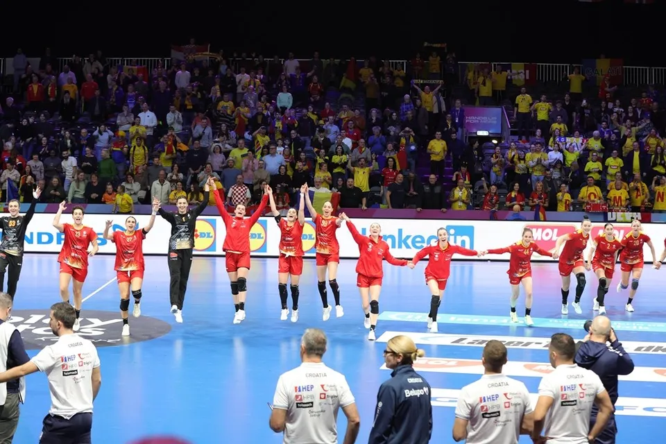 O echipă de handbal feminin în echipamente roșii și negre sărbătorește pe un teren de sport în fața unei tribune pline de spectatori.