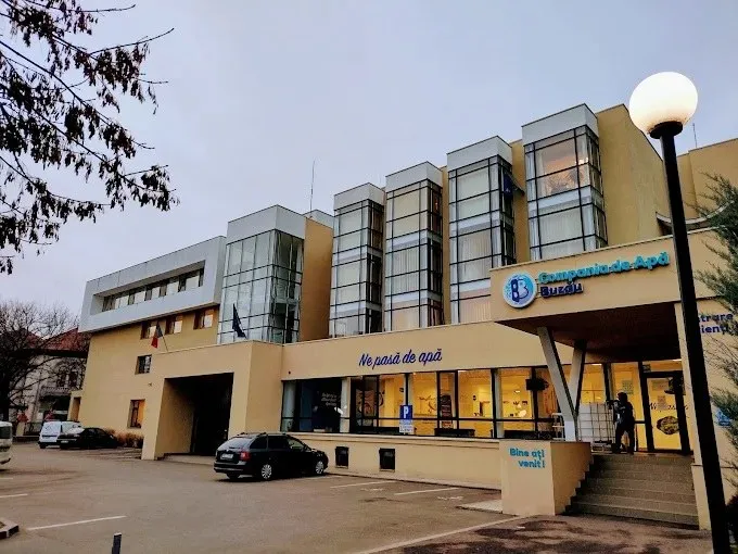 O clădire modernă cu ferestre mari de sticlă, un stâlp de iluminat cu glob alb și câteva mașini parcate în fața acesteia.