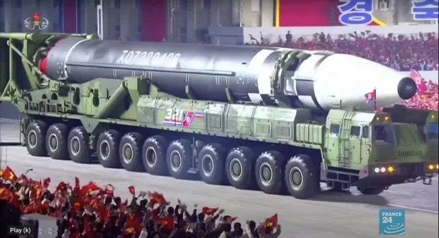 Un rachetă mare de culoare gri și albă este transportată pe un vehicul militar verde cu numeroase roți pe un drum, în fața unei mulțimi.