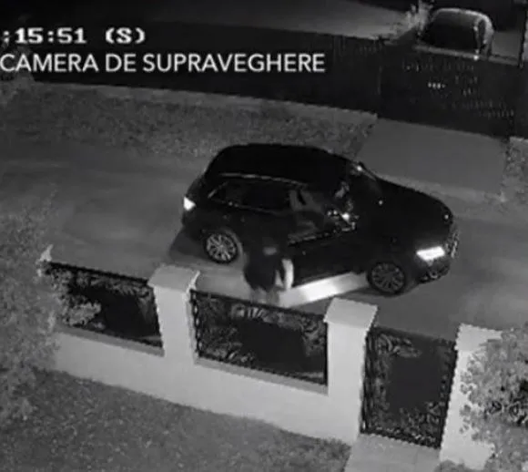O mașină neagră este parcată pe un drum lângă un gard cu porți metalice ornate, surprinsă de o cameră de supraveghere noctuarnă.