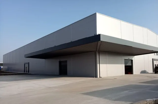 O clădire industrială lungă cu pereți gri și un acoperiș plat dispune de un copertină mare deasupra unei zone de încărcare pe un teren betonat.