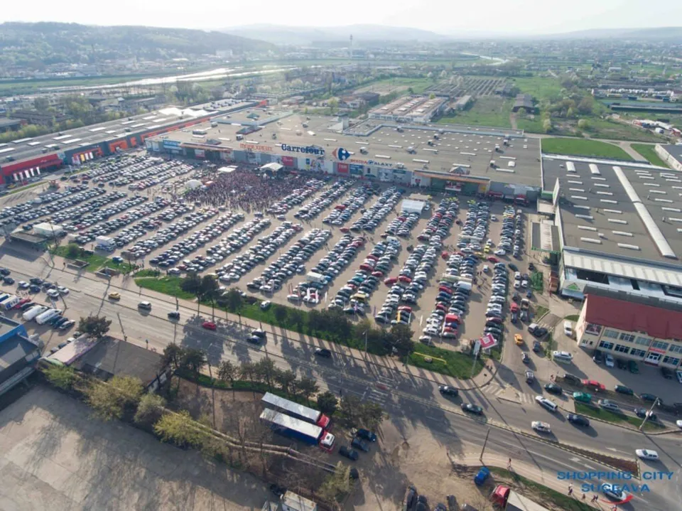 O vedere aeriană prezintă un centru comercial cu un parc de parcare plin de mașini și o stradă cu trafic auto în prim-plan.
