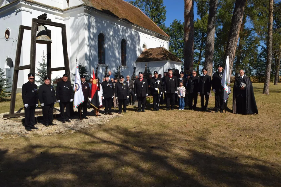 Un grup de persoane în uniforme negre stă în linie pe un teren împădurit în fața unei clădiri albe cu acoperiș de paie.