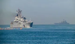 Două nave militare gri navighează pe o suprafață de apă sub un cer senin.